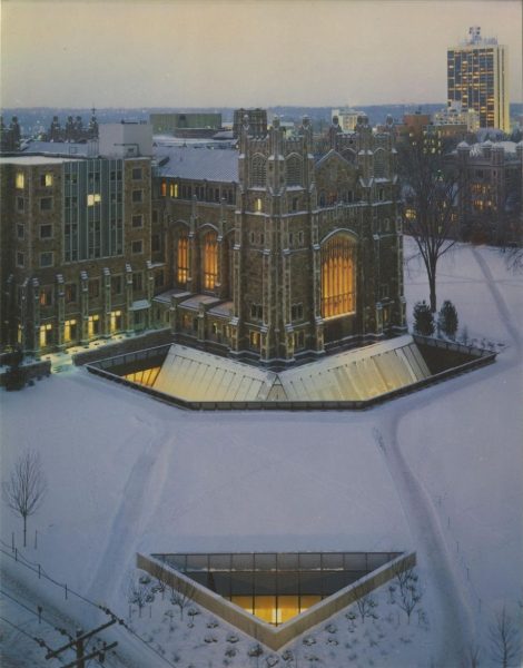 The image portrays the exterior of the University of Michigan Law School during winter. In the foreground, a triangular glass structure is partially visible, set against a snowy landscape.